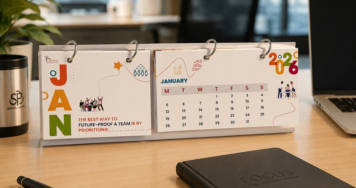 The Catalyst desk calendar displayed on a wooden office desk, featuring a January 2026 page, colourful leadership illustrations, and the message “The best way to future-proof a team is by prioritising.”